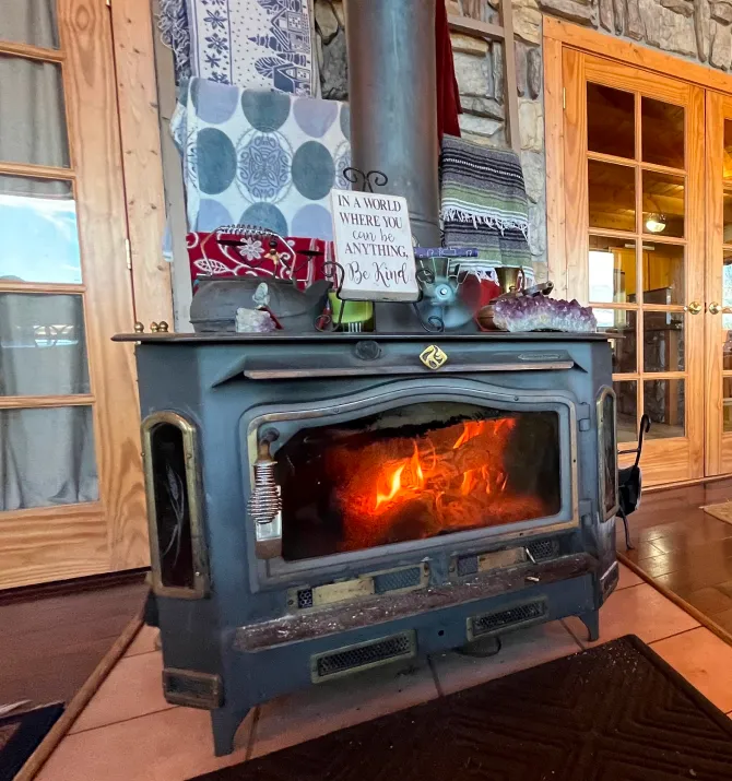 A wood-burning stove with a glass door shows a warm fire inside. A “Be Kind” sign and various items sit on top. The cozy inn room in Pagosa Springs features wooden doors, stone walls, and patterned textiles hanging above the stove.