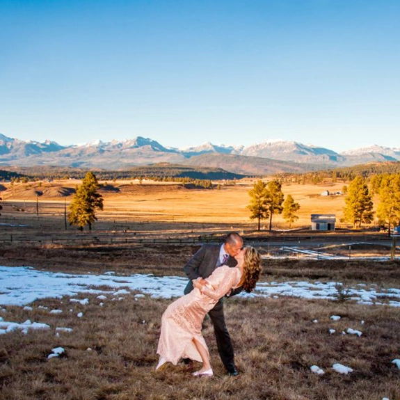 A couple in formal attire poses in a dip, smiling at each other on a snowy field near Pagosa Springs. Behind them, rolling hills, trees, and snow-capped mountains create a stunning backdrop under the clear blue sky.