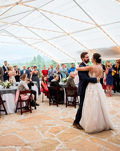 A bride and groom share their first dance under a clear tent decorated with string lights at a charming wedding venue, while guests watch and smile from their tables in the background.