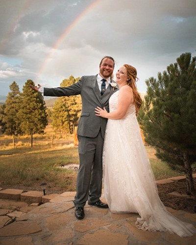A smiling bride and groom in wedding attire stand outdoors on a stone path outside a charming inn, with the groom pointing toward a rainbow in the cloudy sky and trees in the background.