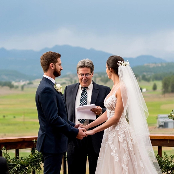 A bride and groom stand holding hands during an outdoor ceremony at the charming inn wedding venue, facing each other as the officiant reads from a paper. Mountains and lush greenery form the scenic background.