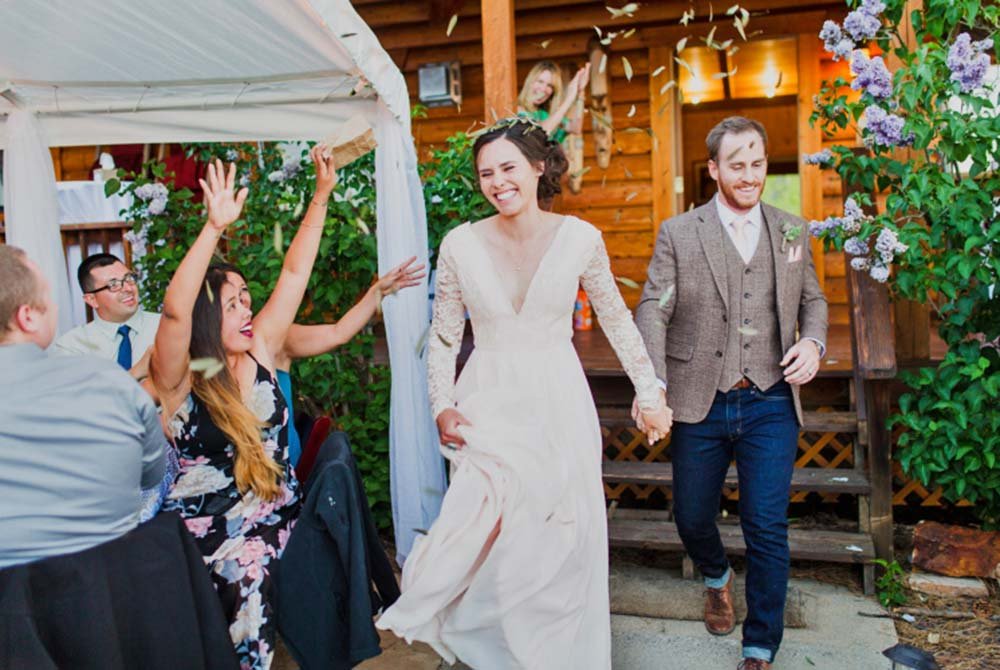 A smiling bride and groom hold hands while walking past excited guests at an outdoor wedding near a charming inn; one guest throws her hands up in celebration. A wooden building and blooming flowers are in the background.