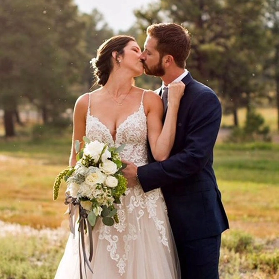 A bride in a white lace wedding dress holding a bouquet kisses a groom in a dark suit outdoors at a charming wedding venue, with trees and sunlight in the background.