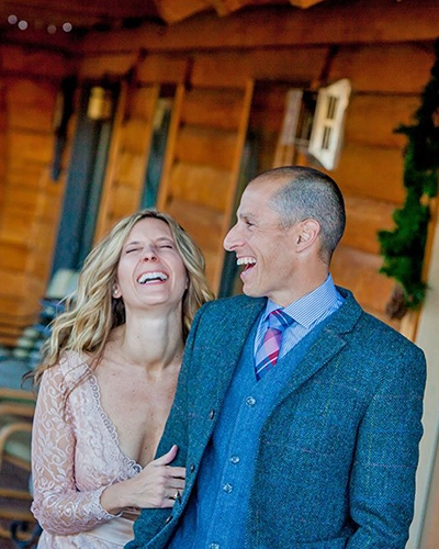 A smiling couple stands together outside a charming inn in Pagosa Springs. The woman, in a light, lacy dress, and the man, in a blue suit with a pink-striped tie, appear to be laughing and enjoying their special lodging experience.